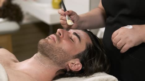 skilled beautician applying a relaxing face mask on a young man
