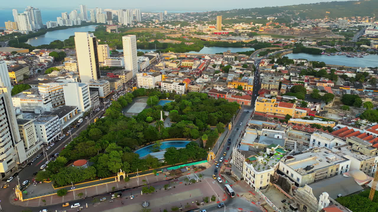 Aerial drone view of Cartagena's city center with a large green park, colonial and modern buildings, and the Caribbean Sea in the background in slow motion