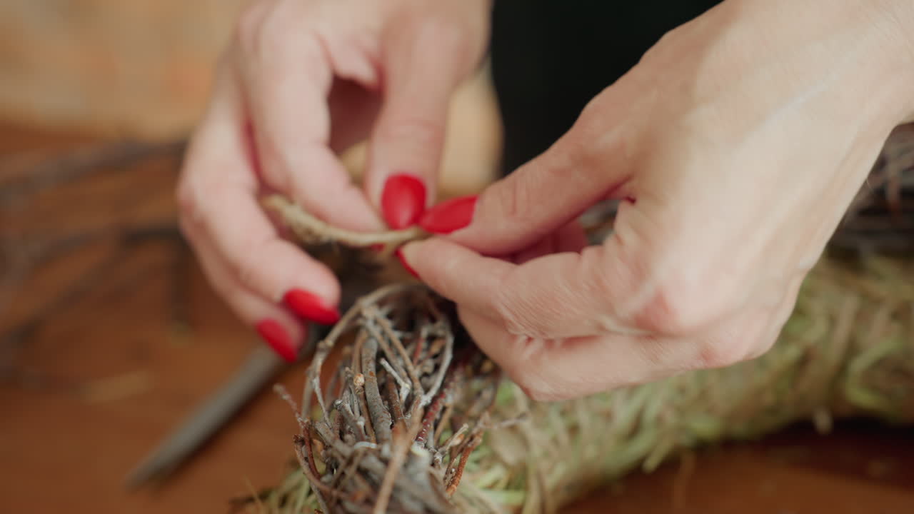 Closeup of florist hands with red nails tying jute rope around handmade wreath of dried grass and twigs on wooden table, securing natural decoration during creative rustic crafting process indoors