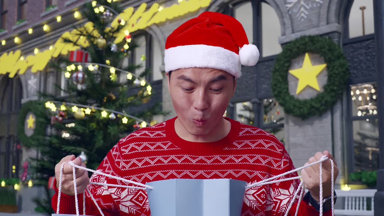 산타 의상을 입은 아시아 남자가 쇼핑 가방을 열고 와우라고 말하고 크리스마스 밤에 서있는 동안 카메라에 미소 짓고 있습니다.