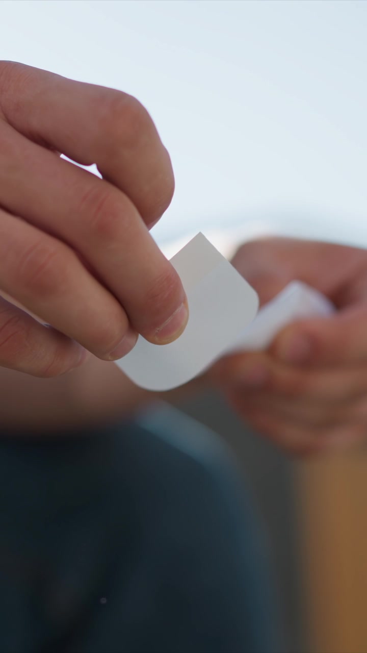 Primo piano delle mani di una persona che maneggia e apre con cura piccoli cerotti adesivi bianchi. L'attenzione rimane sul movimento preciso mentre ogni tampone sterile viene estratto dal suo contenitore, evidenziando