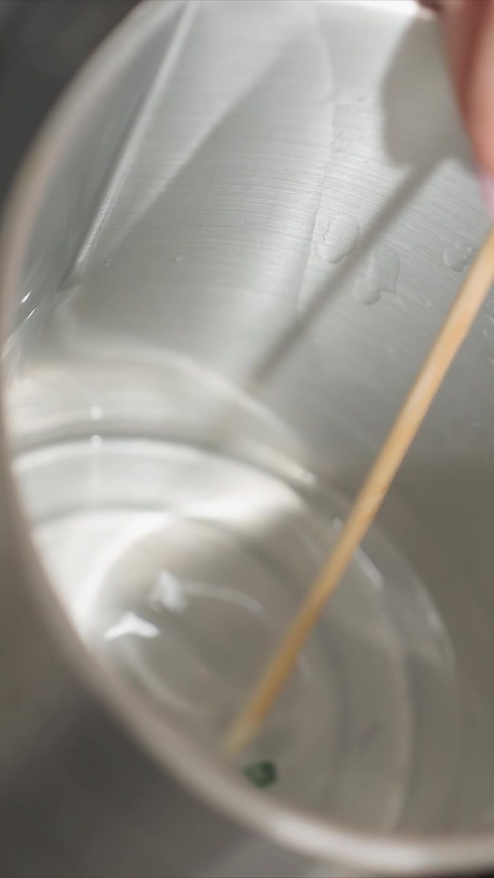 Primo piano di mano che mescola un liquido in un contenitore di metallo con un bastoncino di legno. La mano tiene e muove il mescolatore creando increspature sulla superficie. L'illuminazione soffusa evidenzia i dettagli della texture metallica.