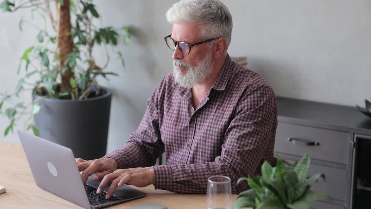 senior businessman in the office is working with a laptop, a gray-haired adult man in a plaid shirt is typing on a laptop or watching a webinar