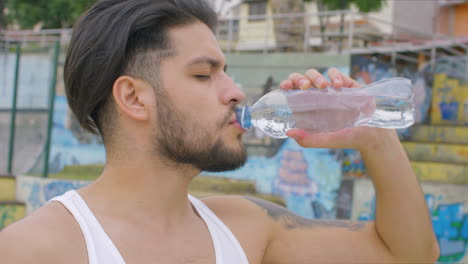 Hübscher-Kerl,-Der-In-Einem-Skatepark-Frisches-Wasser-Aus-Einer-Plastikflasche-Trinkt