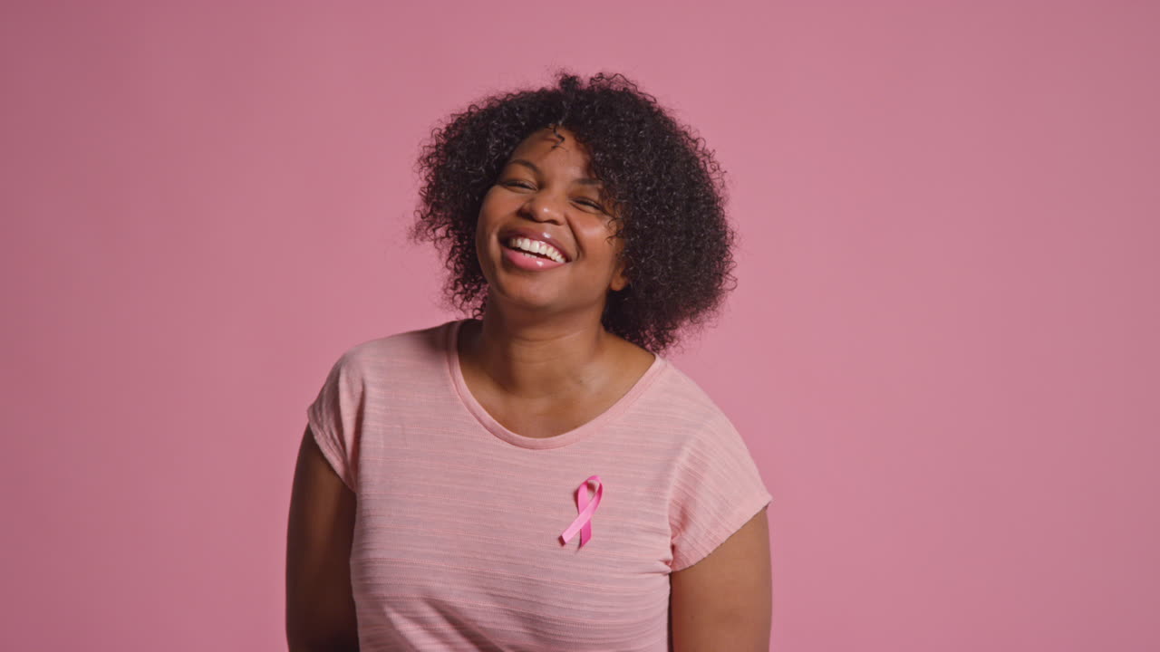 retrato de estudio de una mujer adulta sonriente que lleva con orgullo ropa rosa y una cinta de concienciación sobre el cáncer de mama contra un fondo rosa