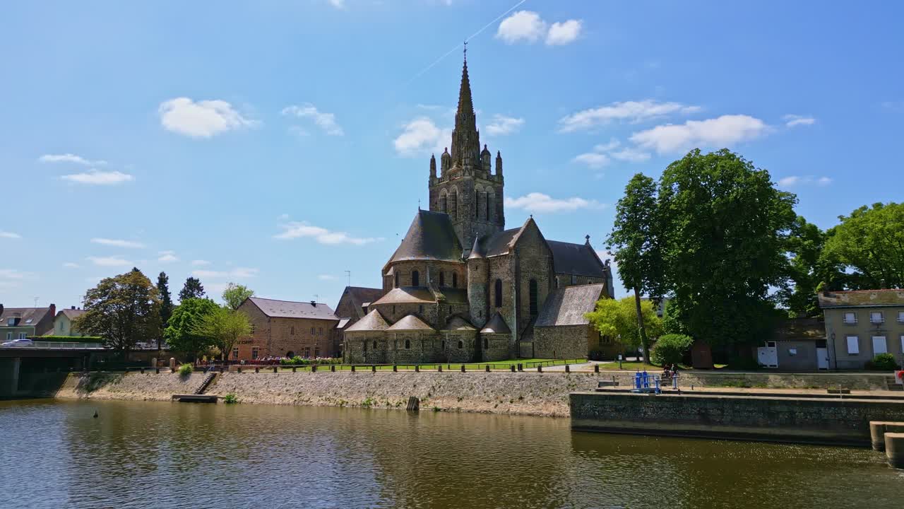 Drone circles Notre-Dame d’Avesnières basilica from Mayenne riverbank; shimmering water and clear blue sky visible.