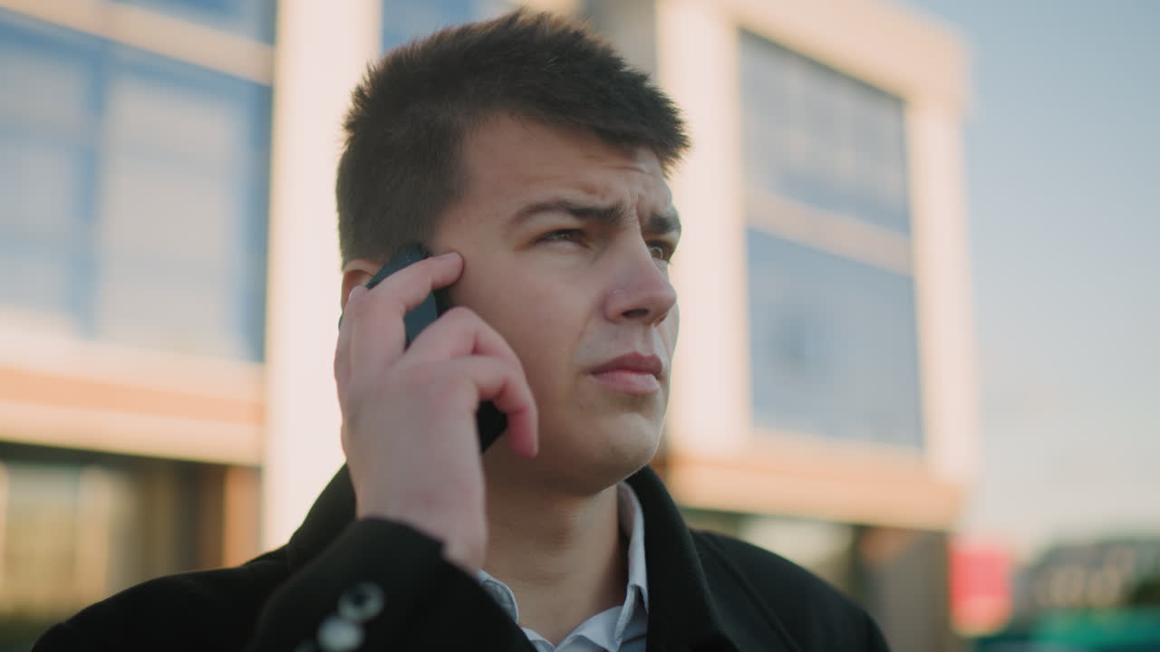 Business tycoon in black suit with white shirt answering phone call outdoors while nodding in agreement, background features modern glass building with soft light