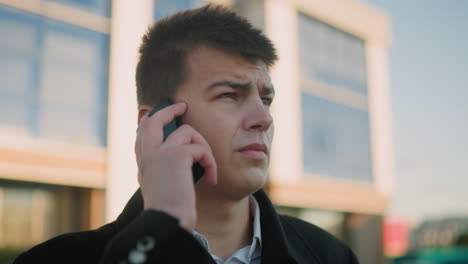 business tycoon in black suit with white shirt answering phone call outdoors while nodding in agreement, background features modern glass building with soft light