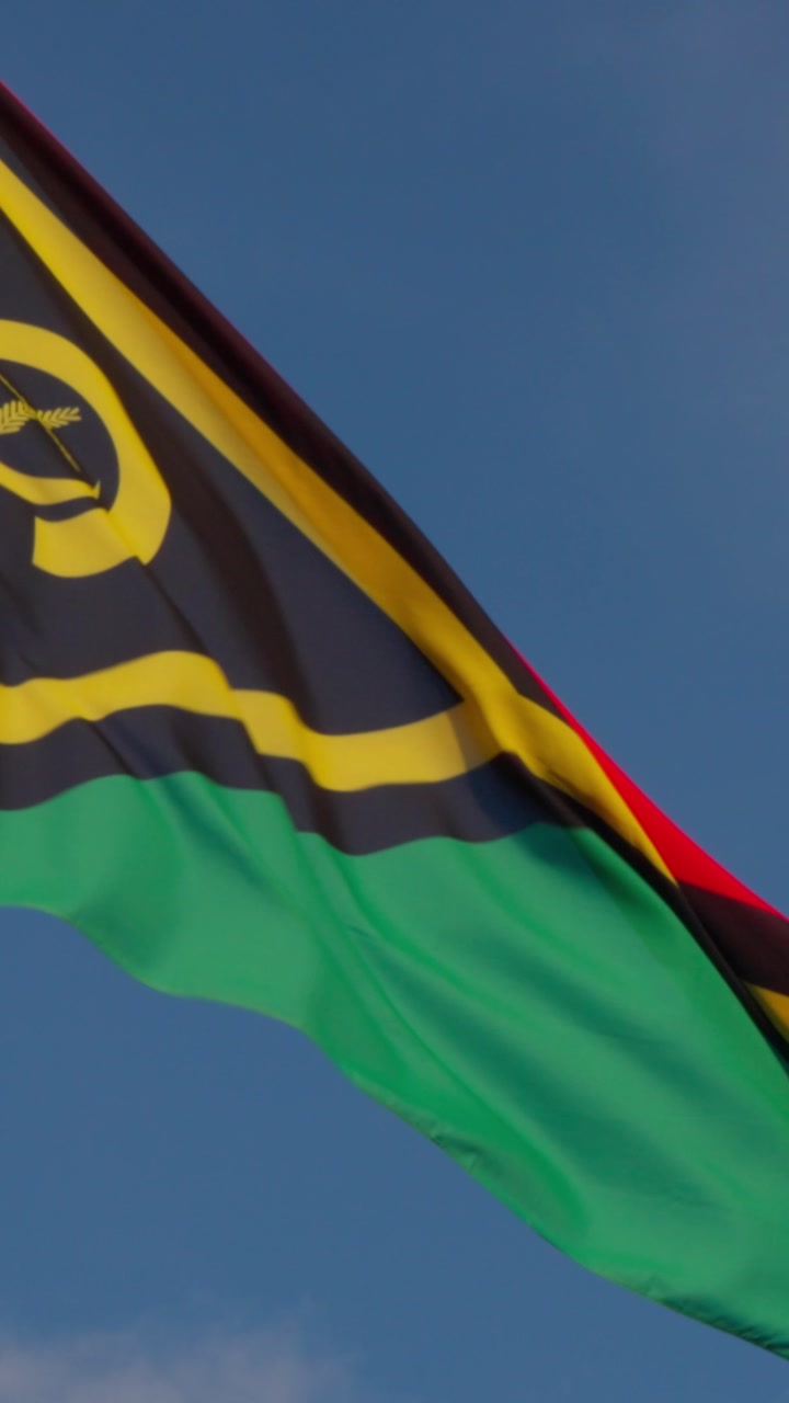Vibrant flag of Vanuatu flutters gracefully in the wind against a clear blue sky. The distinct design features black, red, yellow, and green colors with a boars tusk and fern motif, symbolizing the...