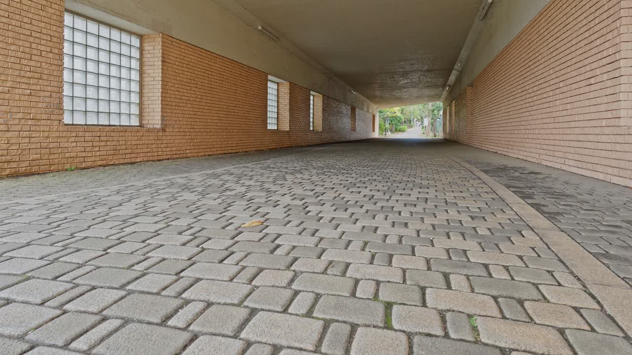 Low-angle shot looking down an empty, straight pedestrian underpass with block paver flooring and brick side walls with glass block windows