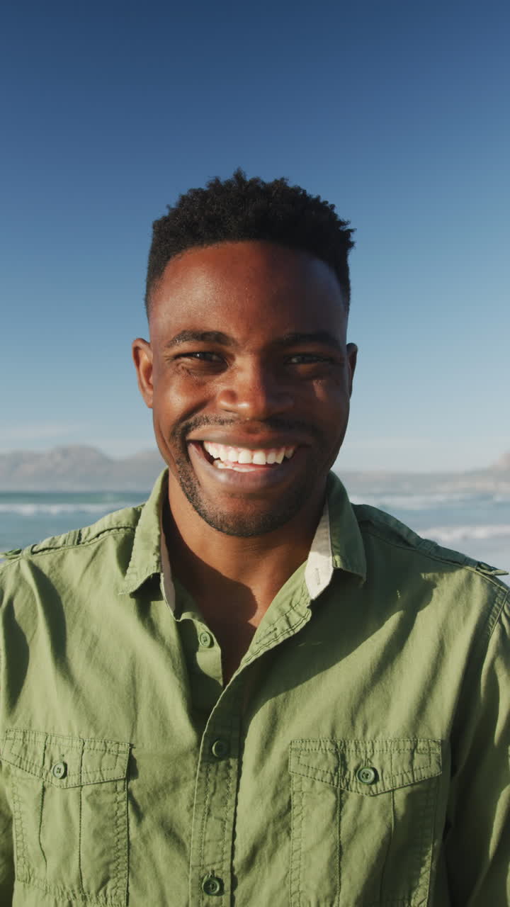 Vertical video of happy arican american man smiling at camera on beach