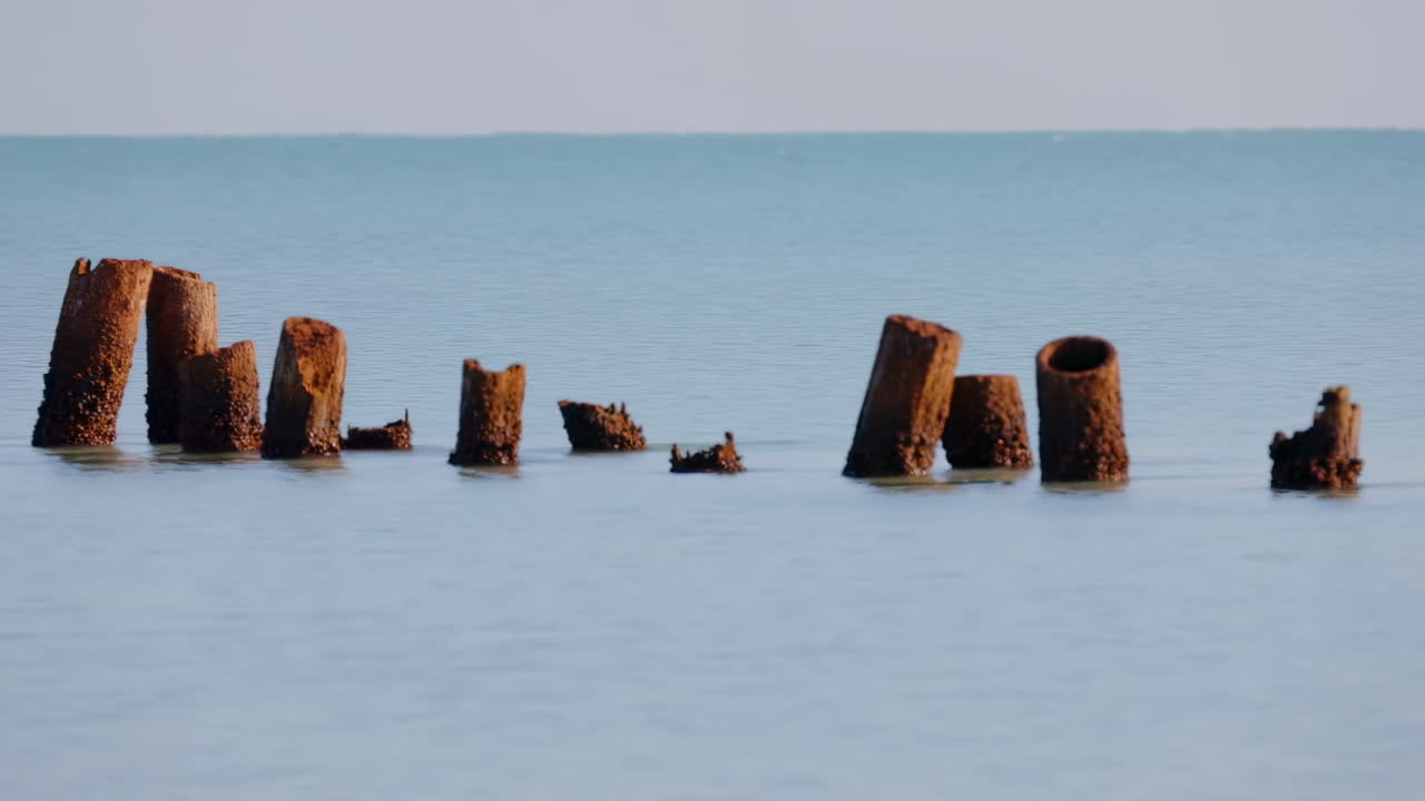 Mare calmo con pali di cemento arrugginiti, pali consumati dal tempo che emergono da acque cristalline, cielo dai colori pastello che si fonde.