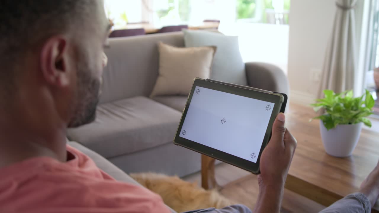 Uomo afroamericano seduto sul divano, inclina il tablet mentre reagisce ai segni negli angoli, indossando una maglietta color corallo.