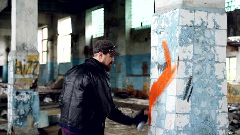 bearded graffiti artist is painting on pillar in old abandoned building using aerosol paint. modern youth subculture, creative people and street art concept.