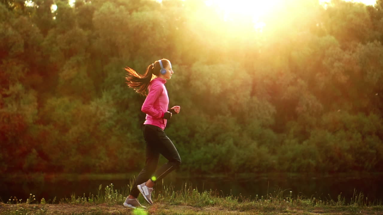A girl in a pink jacket and black pants runs near the river in headphones preparing for the marathon