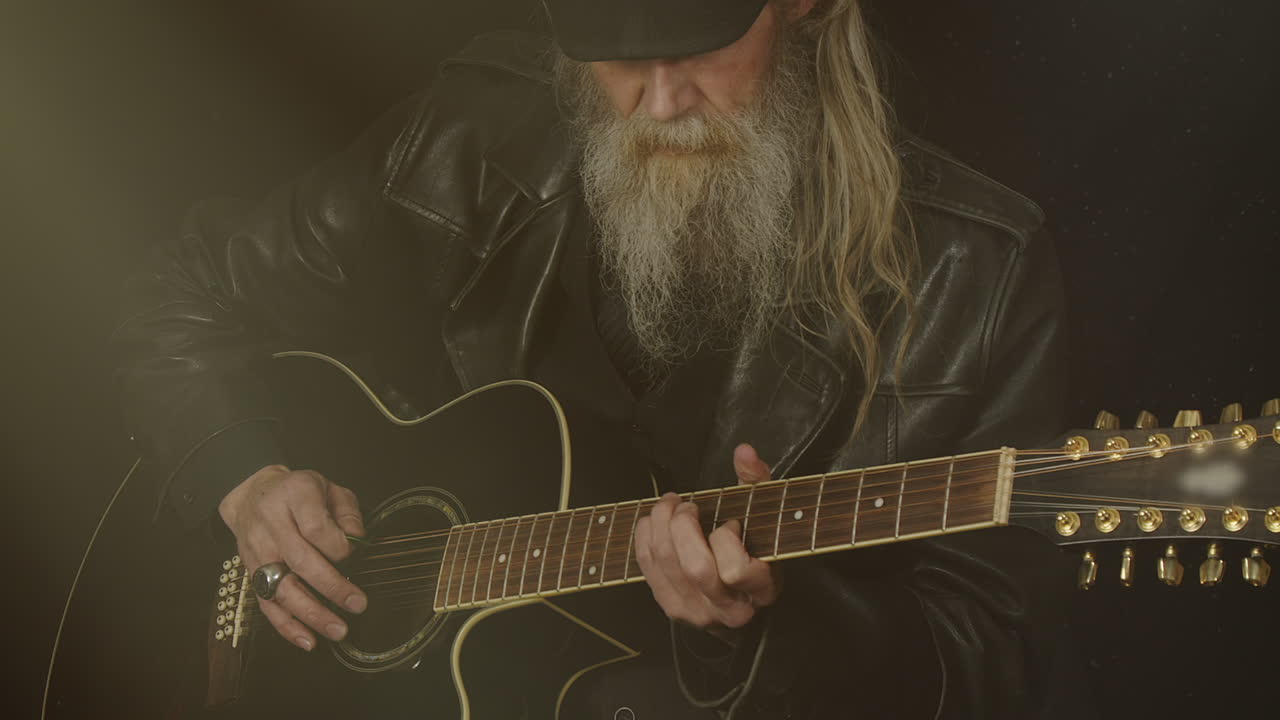 Hip bearded man plays 12-string guitar in smoky dark room, grey hair