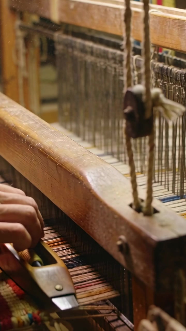 Vertical video: Entering left, hand feeding boat shuttle through warp at workshop loom, weaving rug