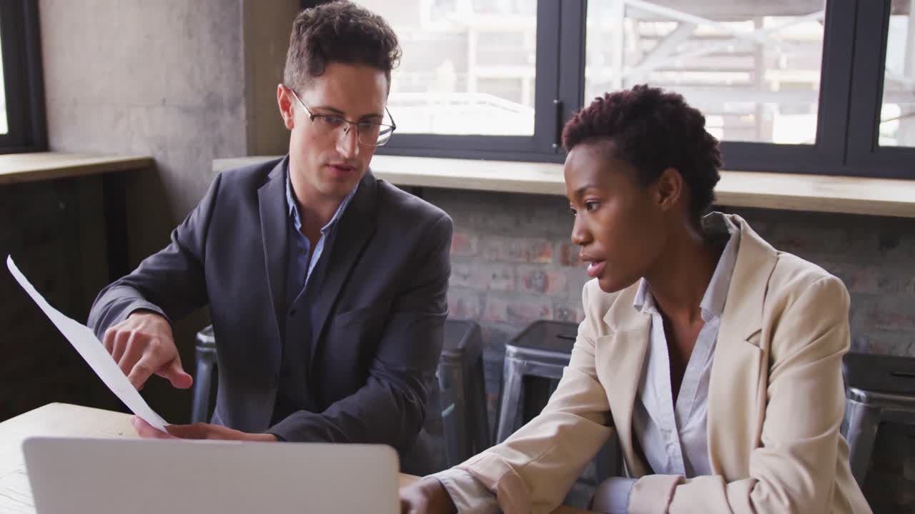 Diverse male and female business colleagues sitting talking in cafe looking at laptop and paperwork