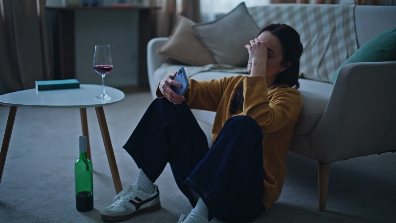 孤独的夜晚，一位压力大的女性坐在地板上，喝着酒看着手机。