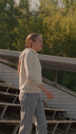 woman walking thoughtfully down stadium steps with relaxed posture, casual hoodie and jeans, background includes rustic stadium seating, electric poles, and lush trees
