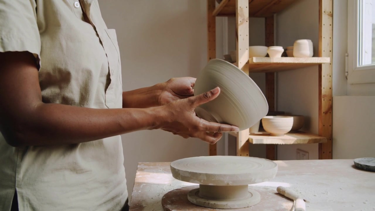 Lifting unglazed bowl from turntable, potter in beige shirt checking rim for symmetry, copy space