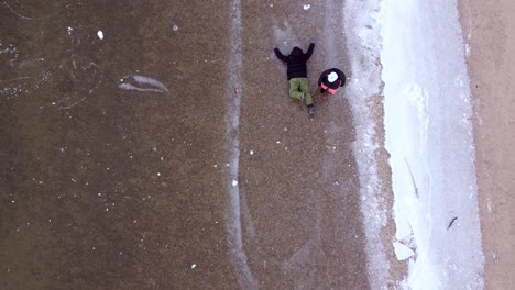 woman dragging man in winter clothing by leg across frozen lake, aerial