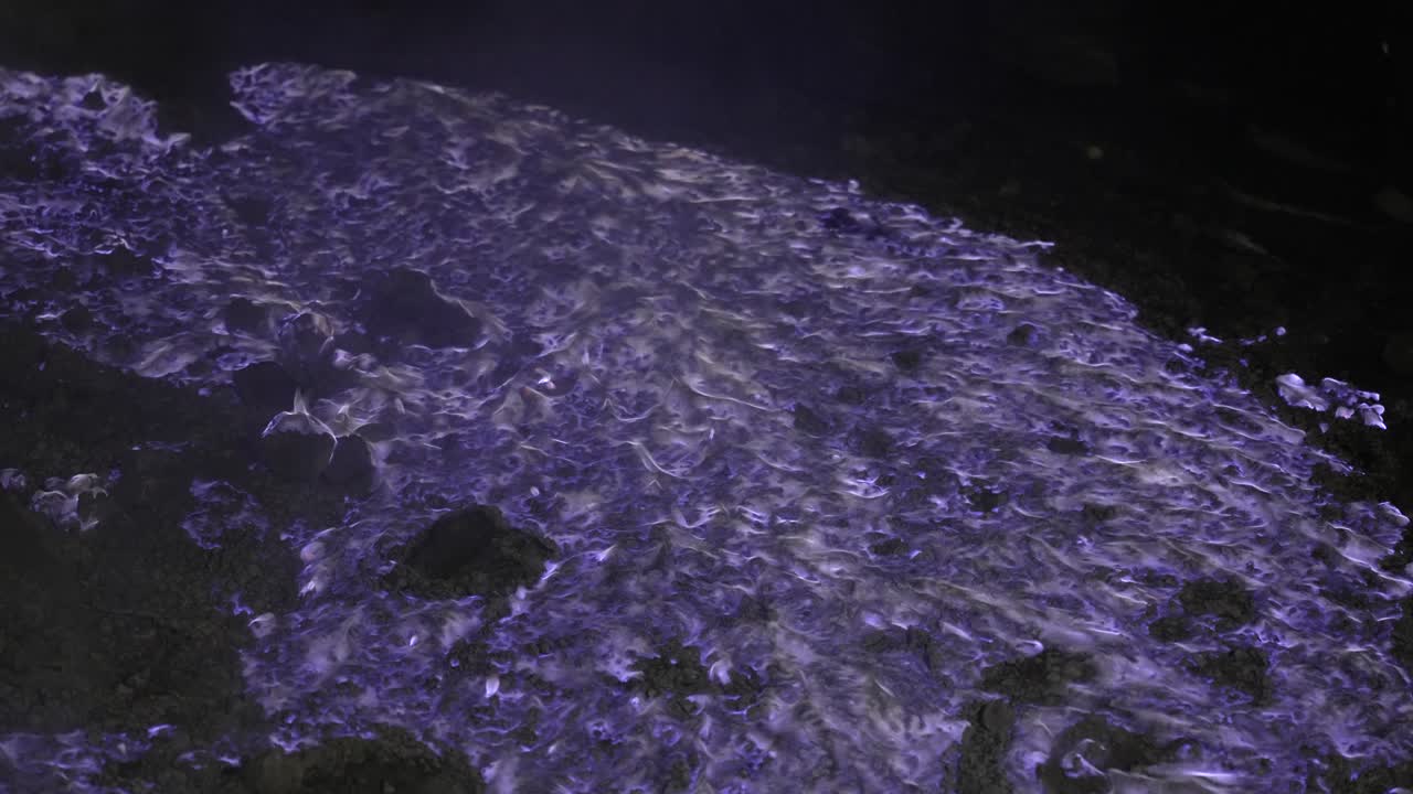 Des flammes bleues de gaz de soufre enflammés scintillent sur un sol volcanique sombre la nuit.