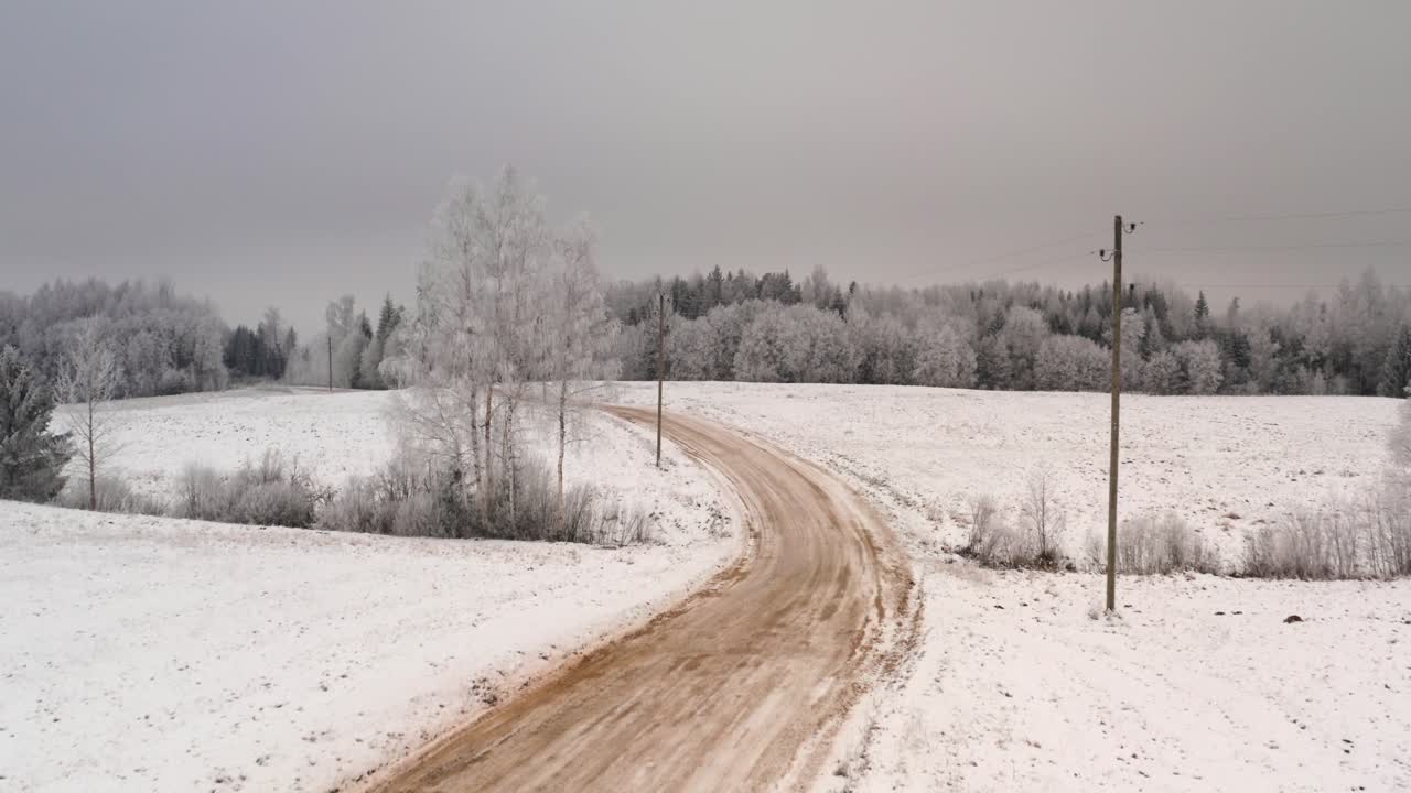 Flying over gravel road in countryside. Aerial winter day scenery with forest covered in snow and frost.