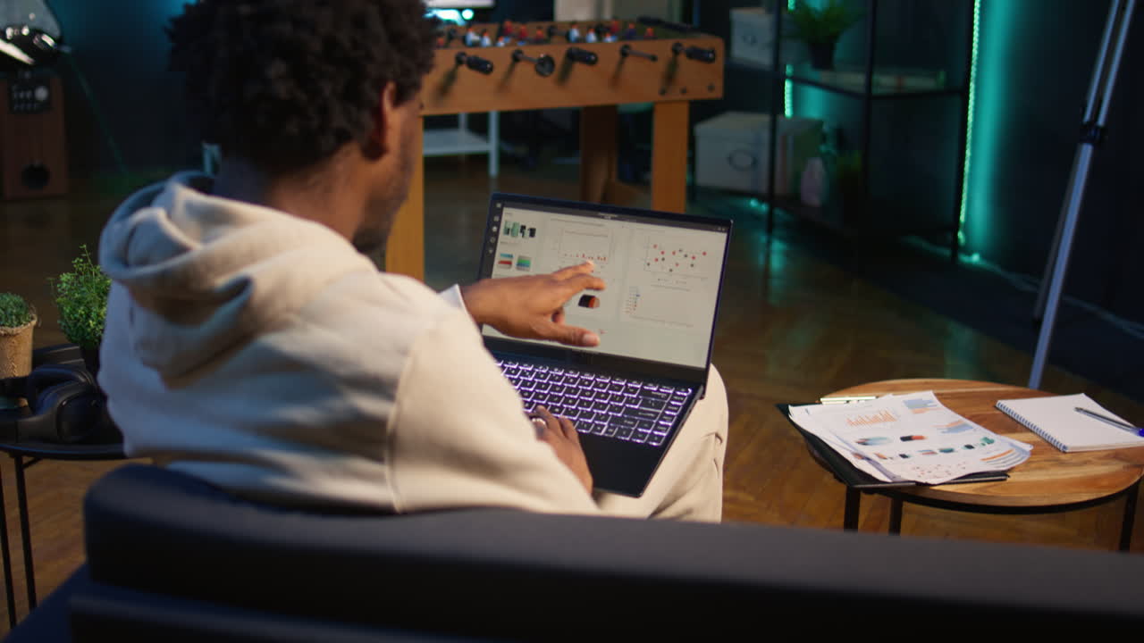 African american person working from home and checking statistical data on laptop screen, analyzing financial graphs. Teleworker in the living room comparing business revenue numbers. Camera B.