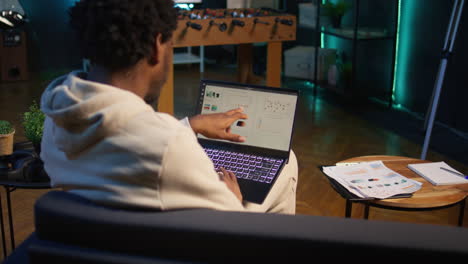 persona afroamericana que trabaja desde casa y revisa datos estadísticos en la pantalla de su portátil, analizando gráficos financieros. teletrabajador en la sala de estar comparando números de ingresos comerciales. cámara b.