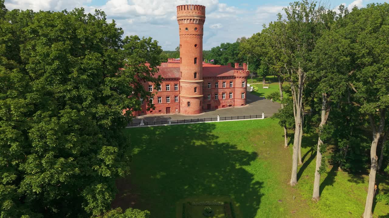 Aerial view of Raudonė Castle in Lithuania, a redbrick medieval fortress with a cylindrical tower surrounded by forest and park on a sunny summer day