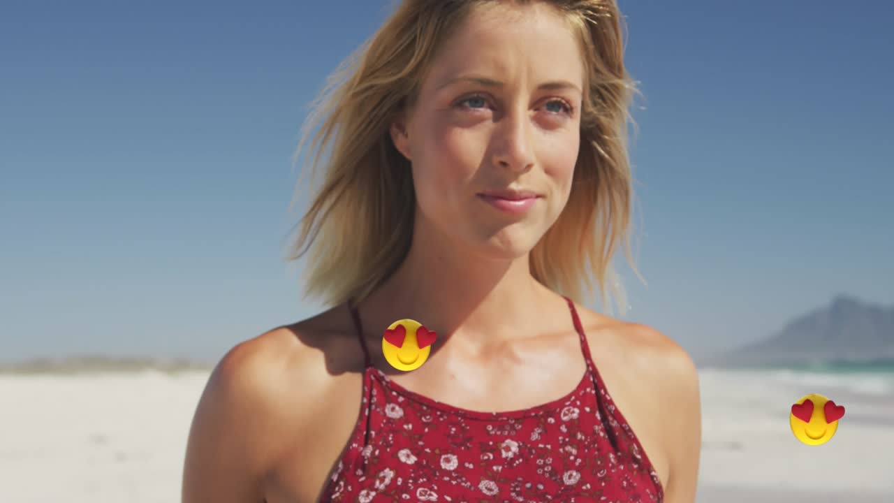Animation af emojis over smilende smuk kaukasisk kvinde, der går på stranden mod himlen