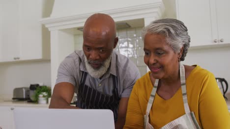 african american senior couple wearing aprons having a video call on laptop in the kitchen at home