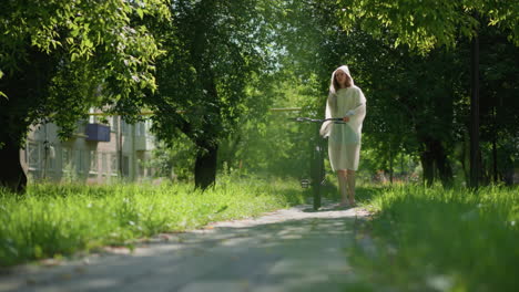 young woman in translucent raincoat walks her bicycle along a sunny pathway, stopping to place the stand, lower her hood, and lean thoughtfully on handlebars, trees, and a building in the background