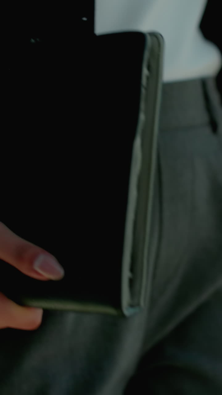 Close up partial view of professional woman holding tablet and coffee cup walking beside iron railing as she sips drink with urban background and parked cars softly blurred by sunlight reflection