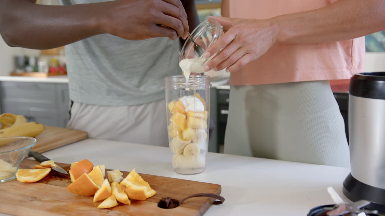 Preparazione di un frullato: coppia multietnica che aggiunge frutta e yogurt nel bicchiere del frullatore in cucina.