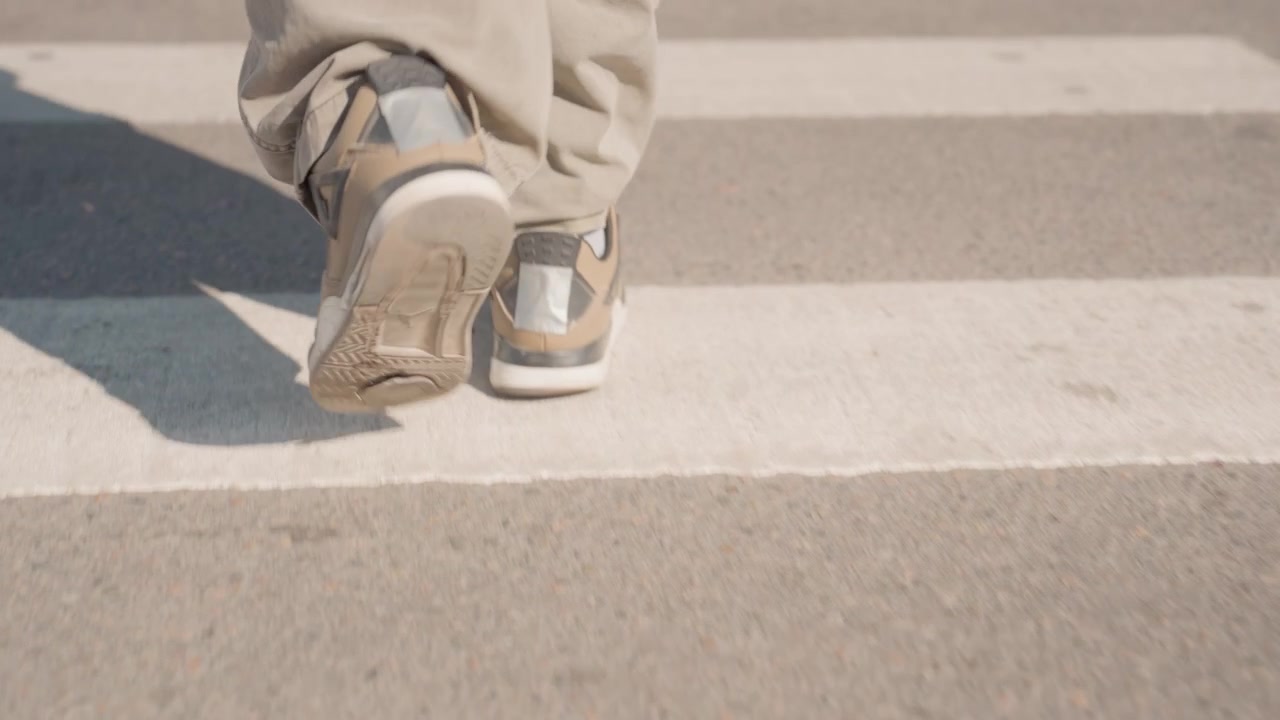 Close up of person feet and legs walking on outdoor crosswalk. subject wears light colored pants and casual sneakers with camera focusing solely on their lower body movement across white