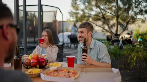 Over-the-shoulder-a-happy-blond-guy-holds-a-bottle-of-beer-in-his-hands-and-actively-communicates-with-his-friends-while-sitting-at-the-table-during-lunch-in-the-courtyard-of-a-country-house