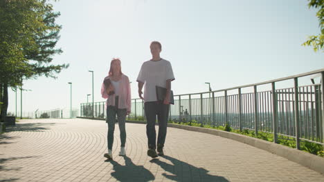 friends walking on interlocked path near railing holding notebooks, engaging in joyful conversation under bright sunlight, surrounded by greenery, cityscape, and warm glow