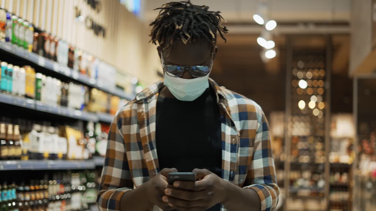 African American man in mask busy with smartphone walks at the store