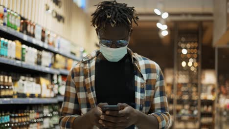 African-American-man-in-mask-busy-with-smartphone-walks-at-the-store