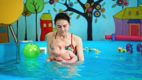 Caucasian brunette woman holds a baby in the swimming pool. Lovely infant wants to get a ball floating on the water.