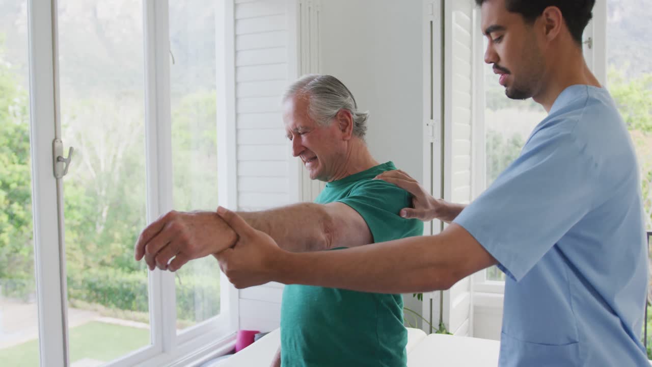 Biracial male physiotherapist slowing moving hand of senior man grimacing with pain at nursing home