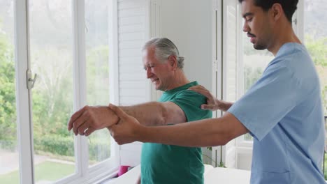 fisioterapeuta masculino biracial ralentizando la mano en movimiento de un anciano haciendo muecas de dolor en un asilo de ancianos