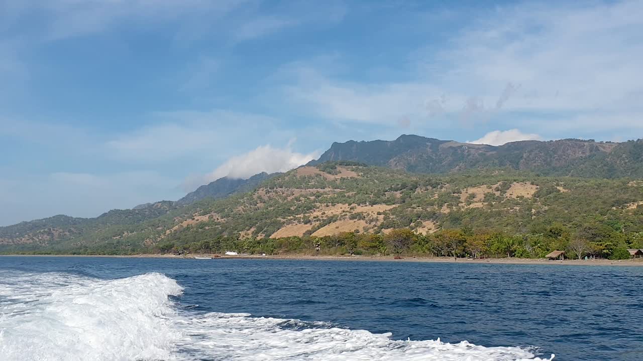Rugged tree-covered coastline and blue sky of remote tropical island Atauro Island seen from the ocean with boat whitewash wake