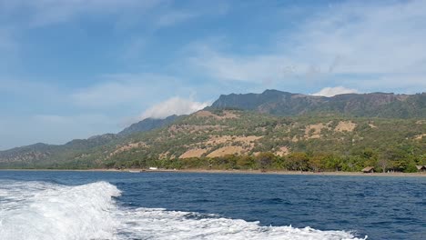 rugged tree-covered coastline and blue sky of remote tropical island atauro island seen from the ocean with boat whitewash wake