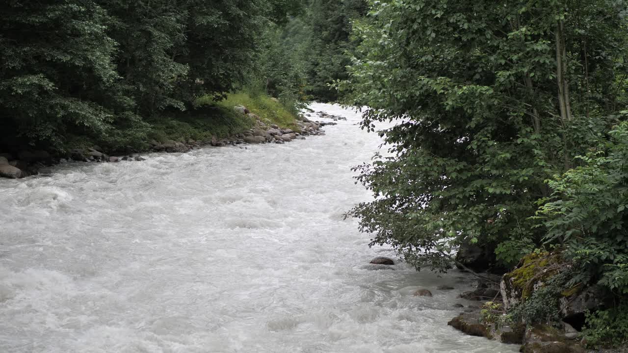 Sungai kecil berisi air abu-abu yang mencair mengalir dan beriak di bebatuan, sementara tanaman hijau dan pepohonan tumbuh di sampingnya. Pengambilan gambar lebar.