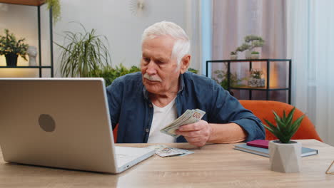happy senior man counting money cash and use laptop pc calculate domestic income earnings at home