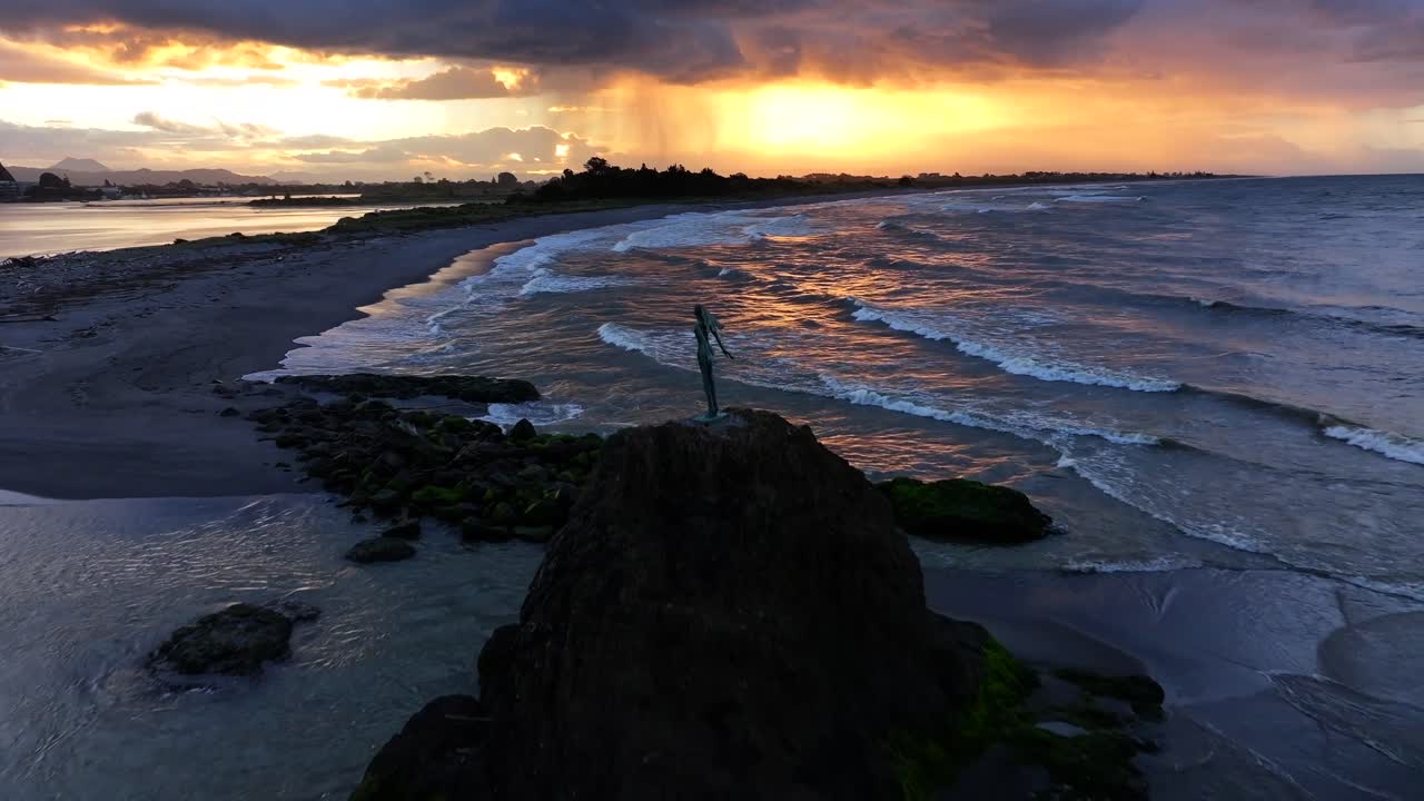 Sunset waves crash against a rocky outcrop, spray rising around a lone statue silhouetted in gold—still and solemn amid the ocean’s restless glow.
