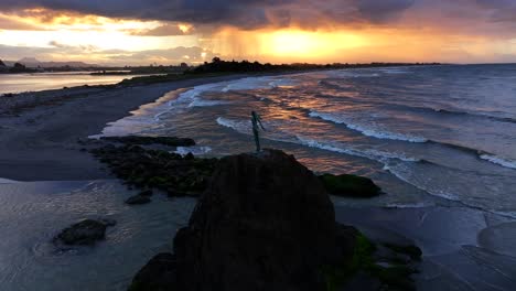 sunset waves crash against a rocky outcrop, spray rising around a lone statue silhouetted in gold—still and solemn amid the ocean’s restless glow.